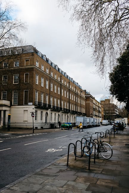 A multi-storey brick building with white window frames and small balconies on the upper floors is situated along a city street. On the pavement directly in front of the building, there are several bic