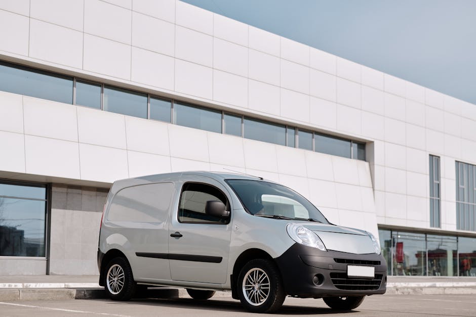 Photograph of a light grey commercial van parked on a paved street in front of a modern white building with large tinted windows. The van has black side mirrors, black front and rear bumpers, and a sliding side door. The vehicle's wheels feature multi-spoke rims, and the van appears to be recently loaded or ready for a home relocation or furniture transport service. The environment is lit with natural daylight under a cloudy sky, indicating daytime conditions. The setting suggests an urban area, possibly during a house removal or move, with the van positioned close to a curb. The company Man with Van Belsize Park is known for providing removals, which aligns with this scene of loading or transportation activity related to packing and moving services.