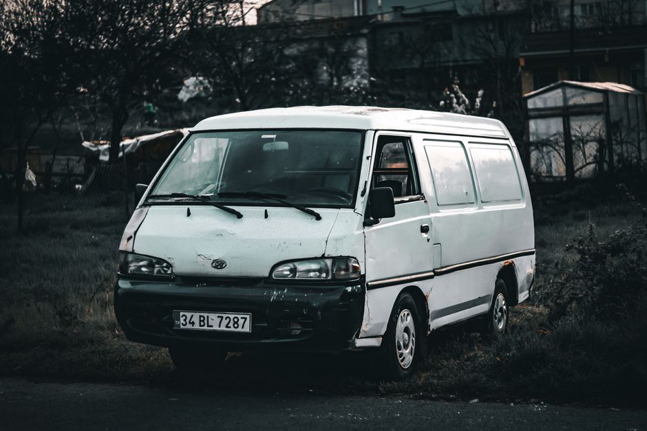 A white commercial van parked on a grassy area next to a residential property, with visible signs of wear such as rust and dents on the front and side panels. The van has a sliding side door, closed windows, black side mirrors, and a black front bumper. The vehicle is situated in front of a house with a garden, with additional trees and structures visible in the background. The scene appears to be taken in low light or overcast conditions, with a slight reflection on the windshield. Outside the van, no moving equipment or furniture is visible, indicating that the vehicle may be parked before or after a home relocation process. This image relates to the services offered by [COMPANY_NAME], such as removals or furniture transport, highlighting the importance of vehicle preparation and careful handling during packing and loading for house moves. As part of the loading process, furniture and boxes would typically be loaded into such a van for transportation to a new address, though they are not present in this specific scene.